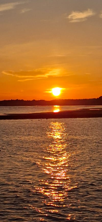 The image shows a sunset over a body of water, likely the Pärnu Bay, in Pärnu linn, Estonia. The sun, a bright orange orb, is partially obscured by a thin strip of land and a dark tree line in the distance. The sky is a gradient of warm orange and yellow hues, with a few wispy clouds. The water in the foreground reflects the setting sun, creating a shimmering path of light across the surface. Several small dark shapes, possibly birds, are visible on the water and the sandbar. The overall scene conveys a sense of tranquility and natural beauty.