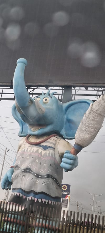 This is a vertical, medium-shot, eye-level, full-color photograph of a large, blue elephant statue in Magodo, Nigeria. The statue is in the shape of a cartoon elephant with large ears, a trunk raised in the air, and a long, thin, bristly object held in its right hand. The elephant is wearing a gray, patterned sweater with geometric shapes and a red necklace. The background is an overcast sky with some blurred lights visible. A billboard for "Eko Oil" is visible behind the statue. The statue appears to be located outdoors, possibly at a roadside attraction or gas station. There are no people visible in the image.