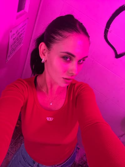 A young adult female is depicted in a close-up selfie taken within an indoor environment characterized by strong magenta lighting. She has dark hair pulled back, blue eyes, and is looking directly at the camera. She wears a red, long-sleeved, ribbed top featuring a small, light-colored Diesel "D" logo on the left chest. A thin silver-colored chain necklace with a small star-shaped pendant is visible around her neck, and a silver-colored hoop earring adorns her left ear. A portion of blue denim trousers is visible at the bottom of the frame. The background consists of light-colored walls. On the left, a notice with illegible Korean text is affixed to the wall. On the right, a large, dark, abstract mark resembling a partial circle or curved line is present on the wall. This scene is located in 마포구, South Korea.
