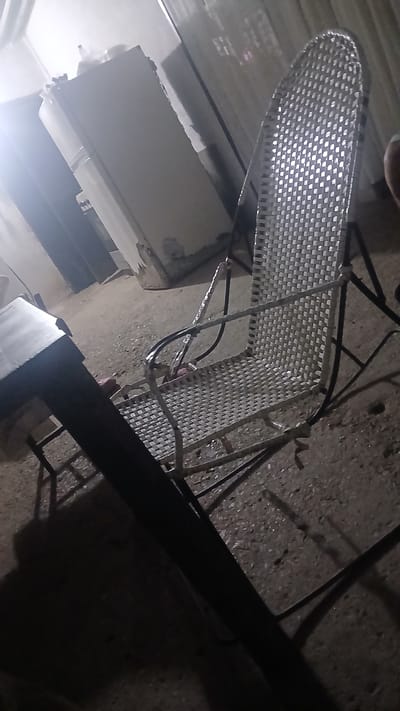 The scene depicts a disorganized, dimly lit outdoor area in Putucual, Venezuela. Dominating the foreground is a metal chair with a woven seat and back, positioned at an oblique angle. The ground is a rough, possibly concrete, surface. In the background, partially obscured by glare, are portions of a white or light-colored building. A thick, dark object, possibly a pipe or beam, crosses the frame diagonally, further contributing to the obscured view. No people or other distinct objects are identifiable. The overall impression is of a cluttered, unkempt exterior space.