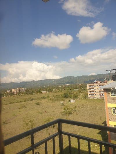 The image presents an elevated panoramic view of a semi-urban landscape in Mumbuni North ward, Kenya, under a bright blue sky with scattered cumulus clouds. In the immediate foreground, a dark metal balcony railing, composed of horizontal and vertical bars, extends across the lower portion of the frame. To the right, a section of a building facade, painted yellow with brown trim and featuring white-framed windows, is visible.
The midground is dominated by an expansive, uncultivated field covered in dry, light-green grass and interspersed with small, sparse bushes. A faint dirt path traverses horizontally across the left side of this field. A lone individual, distinguishable by a red garment, is situated centrally in the field, moving from left to right. Towards the middle-right of the field, a small, light-colored rectangular structure, possibly a shed or enclosure, is visible. Scattered wooden posts, suggesting fencing, are present throughout the field.
In the further midground, multiple multi-story buildings are observable. On the far left, a light-colored building, potentially under construction or recently completed, stands. On the far right, a completed multi-story residential building with light-colored walls and numerous windows features several black water tanks on its roof. The background is defined by a range of green, vegetated hills extending across the entire horizon.