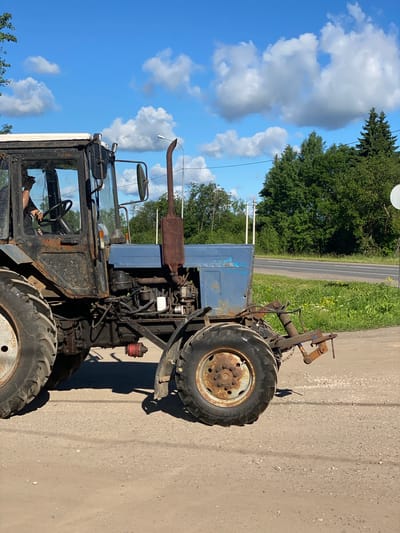 A blue and black tractor, weathered and rusted, is parked on a dirt road in Kaykino, Russia. The tractor is on the right side of the image, facing towards the right. Its large, worn tires are visible, with the rear tire on the right showing a rusty rim. A person is visible inside the driver's cab, with their left arm resting on the steering wheel and holding what appears to be a cigarette. The cab is dark and appears to have some damage or wear. The tractor has a prominent, rusty exhaust pipe extending upwards from the engine block. In the background, a paved road runs horizontally across the image, with a grassy verge and trees lining the far side of the road. The sky is bright blue with scattered white clouds. A white, circular object, possibly a sign or satellite dish, is visible on a pole on the right side of the image. Utility poles and wires are also present in the background. The overall environment suggests a rural or semi-rural setting on a sunny day.