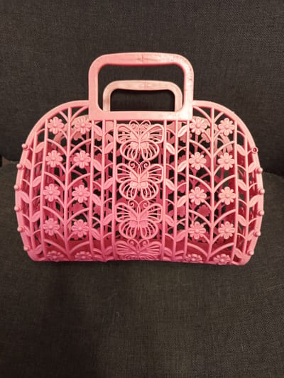 A pink, plastic basket with a cut-out design of flowers and butterflies sits on a dark gray, textured surface, likely upholstery. The basket has a curved, open top with two handles. The intricate design features vertical slats interspersed with delicate flowers and leaves. Three prominent butterfly motifs are centrally located on one side of the basket. The lighting suggests an indoor setting, with no indication of time of day or weather. No people are visible in the image, and there is no visible text. The overall impression is that of a decorative or toy accessory.