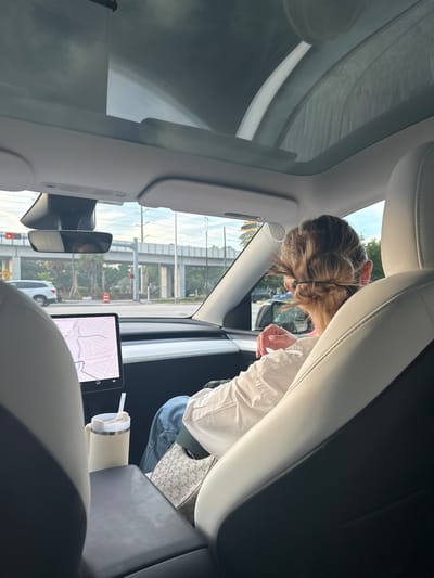 The image shows the interior of a car, likely a Tesla, with a woman's back to the camera. She has her hair pulled up in a bun and is wearing a white shirt and jeans. She is holding a beige travel mug with a straw. A car's navigation system screen is visible in the foreground, displaying a map of an urban area. Outside the car, palm trees and an overpass are visible, suggesting an outdoor setting in a city. The scene appears to be captured during daylight. The presence of a Tesla and the navigation system suggest it is a modern vehicle. The overpass and the type of vegetation hint at a location like Florida.