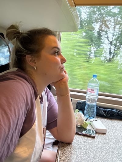 A woman with blonde hair pulled into a bun is looking out a train window. She is wearing a purple and white shirt and has her hand resting on her chin. On the table in front of her are a bottle of water, a mobile phone, a small fan, and a power bank. Outside the window, trees and greenery are visible, suggesting a rural or suburban environment. The motion blur of the scenery indicates the train is in transit.