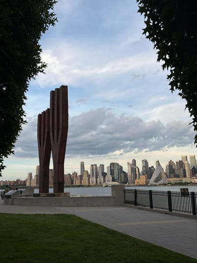 The image shows the "Empty Sky" 9/11 memorial in Weehawken, New Jersey, USA, with the Manhattan skyline in the background. The memorial consists of two tall, rust-colored steel structures that stand against a cloudy sky. The foreground features a manicured green lawn, a paved walkway, and a black metal fence. In the mid-ground, the memorial is situated on a concrete platform. Several people are visible near the memorial, standing and looking towards the Manhattan skyline. The skyline itself is a dense collection of modern skyscrapers, with various architectural styles and heights. A distinct triangular building with a glass facade is prominent among the structures. The overall scene captures a somber memorial site with the bustling city as a backdrop.