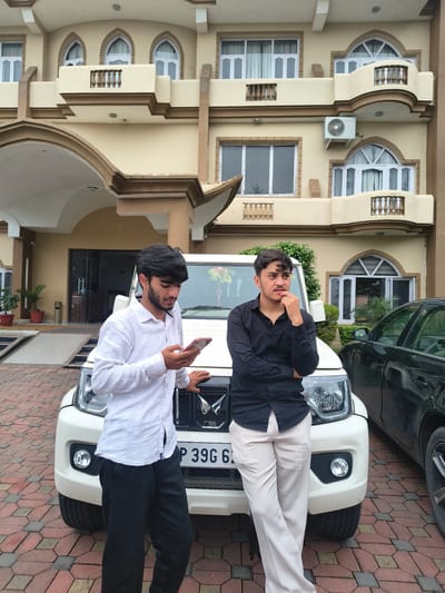In Khaniara, India, two young men stand in front of a cream-colored building. A white SUV is parked directly in front of them. The man on the left, wearing a white shirt and black pants, is looking down at his phone. The man on the right, in a black shirt and light-colored pants, has his hand on his chin, looking to the right. The building behind them has multiple windows and balconies, and an air conditioning unit is visible on the exterior wall. The ground is paved with interlocking bricks. A black car is partially visible to the right of the white SUV.