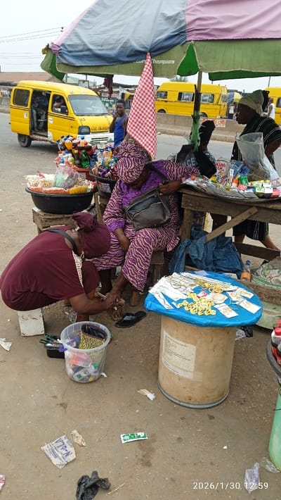 This image, captured on January 30, 2026, at 12:59 PM, depicts a vibrant roadside market scene in Ojokoro, Nigeria. The central focus is a foot care service being performed under a large, multi-colored market umbrella.
A woman, dressed in a purple patterned traditional outfit with a matching headwrap, sits on a small wooden stool, her left foot elevated on a wooden block. She carries a dark shoulder bag. Kneeling before her on a white metal stool, a second woman, wearing a maroon dress and matching headwrap, meticulously attends to the first woman's foot, utilizing tools from a clear plastic bucket positioned nearby.
Surrounding this activity are makeshift market stalls. To the left, a display of packaged snacks and bottled beverages is arranged in a large black basin on a wooden stand. To the right, a wooden table holds various small packaged goods, and an upright wooden cable spool, repurposed as a display surface, exhibits numerous gold-colored jewelry pieces, including necklaces and earrings, laid out on a blue plastic sheet. A third woman, in a striped top and green headwrap, stands near the jewelry display. Further back, a man in a blue tank top stands by the roadside.
The background features a paved road populated by multiple yellow "danfo" mini-buses, common public transport vehicles in Nigeria. One such bus is partially visible to the left with its door open and passengers inside, while others are further down the street. The visible bus has "USE OLORUN" written on its front bumper. The sky is overcast. The ground in the immediate foreground consists of dirt and concrete, interspersed with scattered litter, including plastic bags and paper wrappers.