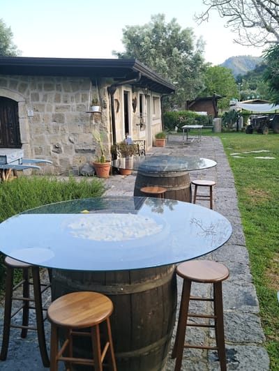 The image depicts an outdoor seating area outside a stone building. In the foreground, a circular glass-top table is supported by a wooden barrel, with three wooden stools arranged around it. The table appears to hold a light-colored, cubed material. Another round glass-top table, also supported by a barrel, is visible in the midground. Behind the tables, a stone building with a dark-tiled roof and windows is partially visible. A pathway made of large stone slabs leads from the tables towards the building. The background features a well-maintained lawn, trees, and a hillside with vegetation, suggesting a rural or semi-rural setting. A tractor is parked on the lawn to the right. The sky is clear and light blue. The scene appears to be a quiet, potentially commercial outdoor space, possibly a restaurant or a vineyard in Pozzuoli, Italy.