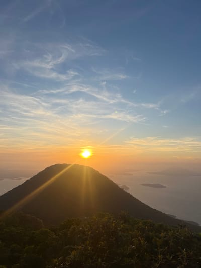 This is a landscape photograph taken from a high vantage point, likely a mountaintop, overlooking a body of water dotted with islands. The sun is setting or rising, casting a warm orange and yellow glow across the sky and reflecting on the water. The sun is positioned just above a silhouetted mountain in the foreground, with its rays fanning out dramatically. The sky is a mix of blue and soft orange hues, with wispy white clouds streaking across it. The foreground is dominated by the dark silhouette of a mountain covered in lush green vegetation. The ocean or large lake stretches out towards the horizon, with several islands of varying sizes visible. The overall atmosphere is serene and picturesque, typical of a scenic natural landscape. The location in El Cacao, El Salvador, suggests this might be a view from a volcanic peak or a high point within the region, known for its natural beauty and coastal proximity.