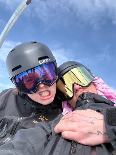 Two individuals are depicted in a close-up, elevated outdoor setting in Le Monêtier-les-Bains, France. The scene is illuminated by a bright sky featuring blue expanse with sparse white clouds. In the upper left quadrant, a metallic gray structural element, consistent with a ski lift component, is partially visible.
The subject on the left wears a dark gray Giro-brand ski helmet and ski goggles with blue and purple mirrored lenses. Their mouth is open, revealing teeth and clear dental aligners. The goggles reflect a distorted image of a face and an indistinct environment. This individual wears a dark gray or black ski jacket with a visible Arc'teryx logo on the chest.
The subject on the right also wears a dark ski helmet and goggles, which have yellow and gold mirrored lenses. A pink patterned bandana is visible beneath their helmet and around their neck. A portion of their face, including a mustache, is visible. Their right hand, adorned with black line tattoos, is positioned on their dark ski jacket. The subjects appear to be in close proximity, suggesting they are seated together.