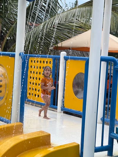 This image depicts a young child within a water park environment located in Juwana, Indonesia. The child, appearing female, stands barefoot on a white, wet ground surface. She is dressed in an orange short-sleeved t-shirt with a white graphic and light pink swim briefs. Water cascades downwards in multiple vertical streams from overhead white support structures. The immediate surroundings include sections of a brightly colored play structure with blue railings and support beams, and yellow panels. One yellow panel exhibits multiple small circular perforations, while another features a large circular cutout. A yellow water slide or ramp is partially visible in the lower-left foreground. In the background, green palm fronds and a beige outdoor umbrella are discernible, with a partially obscured figure in an orange top visible behind the play structure on the right. The child's gaze is directed upward and to the left, towards the falling water.