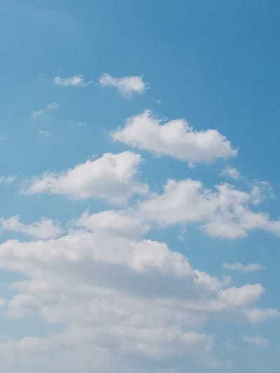The scene depicts a daytime sky over Petroupoli, Greece. The environment consists of an expansive blue sky with scattered white cumulus clouds. Multiple clouds of varying sizes and densities are visible, ranging from small, wispy formations in the upper portion to larger, more defined clusters in the lower and mid-sections. No individuals, man-made objects, or specific ground features are present within the frame. No actions or interactions are occurring.