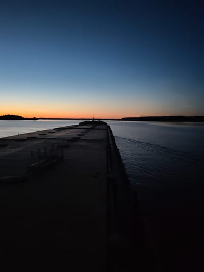 The image captures a dusk scene with a dark, imposing concrete structure extending into a body of water. The sky transitions from a deep blue at the top to a soft orange glow along the horizon, indicating either sunrise or sunset. A faint silhouette of a treeline is visible across the water. The concrete structure, which appears to be a pier or a breakwater, dominates the foreground and midground, casting a long shadow and obscuring much of its surface detail. A few metal railings or supports are visible on the left side of the structure. The water is relatively calm, with faint ripples and a visible wake extending from the right side of the frame, suggesting movement. The overall impression is one of solitude and transition, with the fading light creating a dramatic and somewhat stark atmosphere. There are no people or animals visible in the image. The location is identified as Irdomatka, Russia, though specific landmarks within the city are not discernible from the visual information provided.
