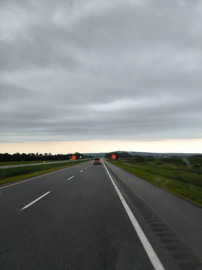 The image shows a highway in Rigaud, Canada, under a cloudy sky. The perspective is from inside a vehicle, looking forward. A red car is visible in the distance, driving on the highway. There are orange construction signs on the side of the road, indicating "TRAVAUX" (construction in French). The landscape features trees and fields on either side of the highway, with hills in the distance. The road has multiple lanes and white dividing lines. The overall impression is of travel on a highway on an overcast day.