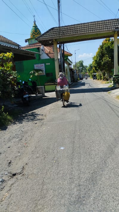 In Kramat Jegu, Indonesia, a woman is seen riding a bicycle equipped with a cart. She is wearing a sun hat and a pink garment covering her upper body, and a mask covering her lower face. Her cart carries several plastic bottles and a yellow bag. The bicycle is on a paved road, which appears to be a street lined with buildings and trees. To the left of the road, there is a green building with a sign that reads "BEKAM" and a mosque with a distinctive green and yellow dome. Several motorcycles are parked near the building. The sky is clear and blue, suggesting a sunny day. The road surface on the left is unpaved and rocky, contrasting with the paved road ahead. In the background, other vehicles and buildings are visible, indicating a populated area.
