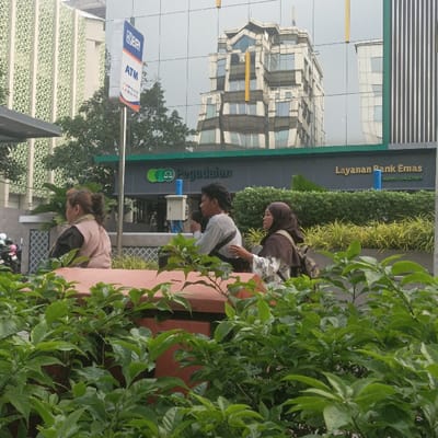 The image captures a daytime urban scene in front of the Pegadaian Cabang Salemba building, located in Salemba, Central Jakarta, Special Capital Region of Jakarta, Indonesia. In the midground, three individuals are partially visible above a reddish-brown planter box and lush green foliage that occupies the foreground.
From left to right, the people include: an individual with brown hair pulled back, wearing a light-colored top, facing right with their back partially towards the viewer; a male individual with dark, curly hair, wearing a light grey long-sleeved shirt and a dark strap across his chest, holding a white object (possibly a microphone or phone) to his mouth as if speaking or singing; and a female individual wearing a brown hijab and a patterned white top with a dark backpack, looking towards the male and appearing to listen.
Behind the individuals, a large reflective glass facade of a building prominently displays the "Pegadaian" logo and name in green, along with "Layanan Bank Emas" (Gold Bank Services) and "CABANG SALEMBA" in white text. To the left, an "ATM BRI" sign is visible on a pole. The glass facade reflects a tall, multi-story building with a complex, multi-faceted architectural design, featuring various window shapes and distinct vertical yellow accents. A building with a white patterned facade is partially visible on the far left. Parked motorbikes are faintly discernible on the lower left. The scene is illuminated by ambient daylight.