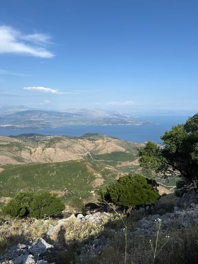 This media file captures a breathtaking panoramic view from a rocky cliffside, overlooking a vast expanse of blue water and a distant, mountainous coastline. The perspective is from high above, suggesting a hike or a viewpoint reached via a strenuous climb. The scene is bathed in bright sunlight, indicating a clear day with excellent visibility. The sky is a vibrant, cloudless blue, with a few wispy white clouds visible on the horizon, adding depth to the distant landscape.

The foreground is dominated by the rugged, grey rock of the cliff face, interspersed with dry, yellowish grasses and a few small, white wildflowers. A large, lush green tree grows precariously from the edge of the cliff, its branches reaching out over the drop. Below the cliff, the terrain transitions to a steep, green, and brown mountainside, with visible tracks or trails winding through the vegetation. The distant view features the shimmering blue water of a large body of water, likely a sea or a large lake, with several islands or peninsulas jutting out from the far shore. The coastline is characterized by layered, hazy mountains, suggesting a significant distance.

The overall impression is one of natural beauty, serenity, and awe. The height and the expansive view evoke a sense of freedom and wonder. The time of day appears to be late morning or early afternoon, given the bright, overhead sunlight and the clear skies. While no people or specific objects are clearly identifiable, the presence of trails hints at human activity, likely hiking or exploring. The location cues, combined with the provided context of Kassopaia Municipal Unit, Greece, suggest a Mediterranean coastal landscape. No visible text is present in the image.