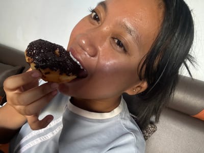 In an indoor setting, a young woman of Southeast Asian descent is captured in a close-up, head-and-shoulders shot, actively biting into a chocolate-frosted donut with dark sprinkles. She is wearing a light blue t-shirt with white piping around the collar and down the front. Her dark hair is visible, framing her face and falling over her shoulder. The woman's face is turned slightly towards the camera, with her right eye appearing more prominent. Her skin has a dewy sheen, possibly from sweat or product. A small, gold stud earring is visible in her right ear. The background consists of a textured, dark brown surface, likely upholstery, and a plain white wall. The scene does not provide any specific visual cues to pinpoint a location within the city of Jambearum, Indonesia.