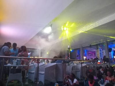 This image depicts a bustling indoor entertainment venue, likely a nightclub or lounge, located in Syokimau, Kenya. The scene is illuminated by diverse artificial lighting, including a prominent disco ball emitting numerous yellow-green light beams and scattered spots across the white ceiling and floor. Haze or smoke is visible throughout the upper midground, contributing to the atmospheric effect.

In the foreground, a metal railing separates the immediate viewing area from the main floor. To the left, a group of four individuals stands conversing around a table where drinks are visible. One person has long dark hair and wears a striped shirt, another is in a light blue shirt. Adjacent to this group, a tall shisha pipe with a blue hose stands upright next to a white wicker-style chair with a cushion.

Throughout the midground and background, numerous patrons, predominantly young adults, are seated and standing at tables furnished with similar white wicker-style chairs. Many are engaged in conversation and appear to be socializing. A partial sign displaying "SIR WALKER" or "VIP WALKER" is discernible in the mid-background. The distant background features architectural structures, possibly a bar or performance stage, highlighted with vivid blue and purple ambient lighting. The overall environment suggests an active and lively social gathering.