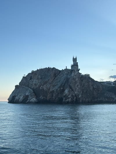 The image depicts Swallow's Nest, a decorative castle located on a cliff near Gaspra, in the Гаспринский поселковый совет region of Ukraine. The castle sits atop a rocky promontory. The sea surrounds the base of the cliff. The sky is a clear, pale blue. The photo is taken from a low angle, with the castle appearing vertically oriented. The castle's architectural details are visible, with its spires and turrets. The sea displays vertical lines, suggesting ripples on the surface.