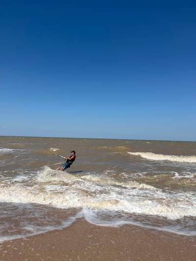 A person is kiteboarding in the murky, brown water of the Sea of Azov off the coast of Dolzhanskaya, Russia, on a clear, sunny day. The kiteboarder, wearing a dark vest, blue and white striped shorts, and a black baseball cap, is leaning back against the pull of the kite, which is out of frame. Water splashes up around the kiteboarder's legs. The waves are small to medium, with whitecaps breaking closer to shore. The beach in the foreground is sandy, and the sky is a clear, bright blue.