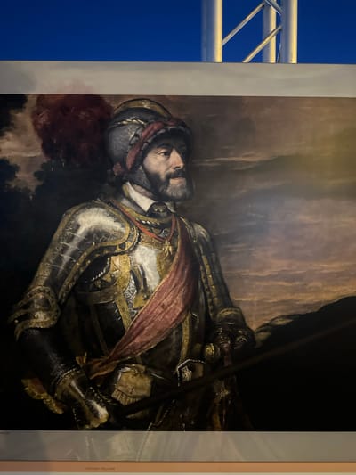 The image is a photograph of a painting displayed in a room. The painting depicts a man in ornate armor, wearing a helmet with a red plume. He has a beard and a red sash across his chest. The background of the painting is a cloudy, atmospheric landscape. Below the painting, a label reads "PINTURA ITALIANA." The room itself is dimly lit, with the top of the frame showing a blue sky and silver metal scaffolding. The overall impression is of an art exhibition. The location in Mazarrón, Spain is not discernible from the visual information provided.