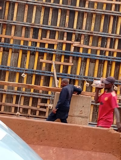 The image shows two men on a construction site in Katambe, Nigeria. The background is dominated by a wooden and metal formwork structure, likely for concrete pouring. The formwork consists of vertical wooden planks painted yellow and dark gray, interspersed with horizontal blue metal bars. Some wooden planks are also visible within the structure.

The subject on the right is a man wearing a red t-shirt with a crest and a black baseball cap. He is carrying a flat object, possibly a piece of wood or material, on his head. The subject on the left is wearing a dark blue long-sleeved shirt and dark pants. He is leaning on a concrete block, looking away from the camera.

The scene suggests ongoing construction work. The lighting indicates it is daytime. The overall atmosphere appears to be one of work and labor. There is no visible text in the image.