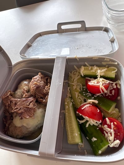 The image shows a close-up, overhead view of an open bento-style lunchbox. The lunchbox is divided into two compartments. The left compartment contains a meat dish, possibly pulled pork or beef, served over what appears to be mashed potatoes or a similar creamy base. The meat is shredded and has a dark brown color, with some lighter, fattier pieces visible. The right compartment contains chopped vegetables, specifically cucumber slices and tomato wedges, all sprinkled with shredded white cheese. The lunchbox lid is open and rests behind the main container. The scene is set on a white, textured surface, possibly a table. In the background, a stack of clear glass containers is visible. The lighting suggests an indoor setting. There are no people or animals present in the image. The overall impression is one of a prepared meal ready to be consumed.