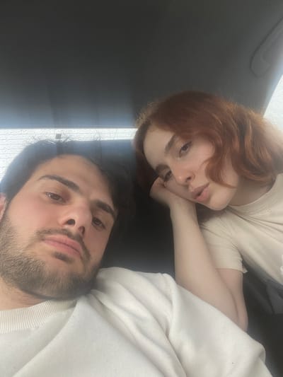 Two people are lying on a dark, possibly black, surface. A woman with red hair and a beige top rests her head on her hand and looks at the camera. A man with dark hair and a beard is next to her, wearing a white sweatshirt. The environment appears to be indoors, with a window showing rain outside. The specific location in Unknown, Georgia, is not discernible from the image.