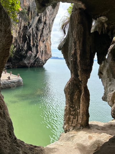 The image shows a view from inside a cave overlooking a body of calm, green water. Jagged rock formations frame the opening of the cave. To the left, a small stone pier with wooden posts extends into the water. In the distance, a large rock formation with vegetation on top rises from the water, and beyond that, islands and the horizon are visible under a cloudy sky. The location is identified as Krasom, Thailand.