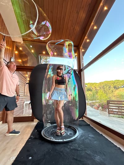 A man, appearing to be in his 50s, wearing a pink polo shirt and dark shorts, stands to the left of the frame, holding a bubble wand. He is looking upwards and to the right, presumably at a large bubble. In the center of the frame, a young woman, wearing sunglasses, a black crop top, and a denim skirt, stands inside a large, iridescent bubble. The bubble is cylindrical and appears to be made of soap film. She is looking forward and smiling. The scene appears to be set on a covered patio or sunroom, with wooden walls and a sloped ceiling. Large windows to the right offer a view of a lush green landscape with trees and potted plants, suggesting a location in Sant Vicenç De Montalt, Spain, known for its Mediterranean climate and natural beauty. The floor is a combination of light-colored wood and a black mat beneath the bubble. String lights are visible on the ceiling, adding to the ambiance.