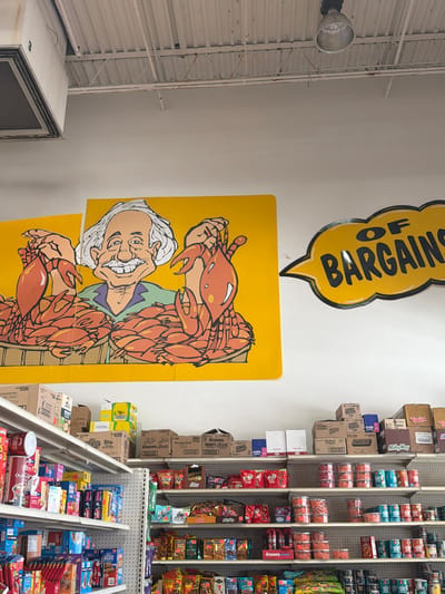 This image depicts the interior of a store, likely a grocery or convenience store, featuring a display of various snack items and a large, colorful mural on the wall. The mural is a cartoonish illustration of an elderly man with white hair and a wide smile, superimposed with a large crab, suggesting a seafood theme. Above the mural, a yellow speech bubble sign reads "OF BARGAIN" in black, bold lettering.
The shelves are stocked with a variety of packaged goods, including candy bars such as Snickers, Milky Way, and Twix, as well as boxes of Hershey's Kisses and other confectionery items. Several boxes are stacked precariously on shelves and on the floor, indicating a busy retail environment or perhaps a sale. The lighting appears to be from overhead fluorescent fixtures, typical of such stores, and the overall atmosphere is bright and utilitarian. The image also shows a large, industrial-looking ventilation unit on the left side, and the walls are painted a light color, creating a stark contrast with the vibrant mural. The time of day is not discernible, but the indoor setting suggests it could be any time. The store's location is indicated as Elizabeth City, United States of America. There are no people visible in the image.