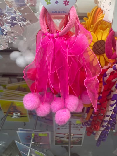 The image shows a close-up of a display of hair accessories, likely in a retail setting. The primary focus is a set of bright pink hair ties with sheer pink tulle fabric hanging down and several fluffy pink pom-poms attached to the bottom. To the right of these, a bright yellow sunflower-shaped hair accessory is visible, followed by other colorful, coiled hair accessories in shades of red, white, and purple. In the background, more hair accessories are displayed, including some with star-shaped charms. The items are suspended and appear to be hanging from a retail display rack. The background is somewhat blurred, but suggests a store environment with shelves and other products. The phrase "MADE IN CHINA" is visible on a tag attached to the pink hair ties. There are no people visible in the image. The overall scene is brightly lit and colorful.