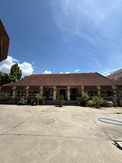 This is a wide-angle, eye-level shot of a traditional Vietnamese house on a sunny day. The house, likely located in Hội Tín, Vietnam, features a red-tiled, pitched roof with exposed wooden beams on the eaves. The facade is light yellow, with a covered porch supported by four fluted columns. The porch is lined with decorative pottery, planters filled with various greenery and flowers, and hanging baskets. A set of stairs leads up to the entrance, which is partially obscured. A large tree with lush foliage is visible to the left of the house, and another building with a similar roofline is seen to the right. The foreground is a concrete courtyard with visible cracks and stains, and a teal-colored garden hose lies coiled on the ground. The sky is bright blue with scattered white clouds. There are no people or animals visible in the frame.