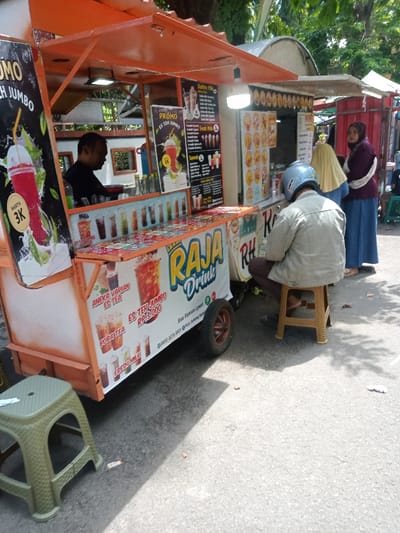 An outdoor scene in Nganjuk, Indonesia, depicts a brightly colored orange mobile beverage stall named "Raja Drink" situated on an asphalt pavement. The stall features prominent advertisements for "Es Teh Jumbo" (Jumbo Iced Tea) at a promotional price of 3,000 Indonesian Rupiah, alongside other beverages like Boba Tea, Fresh Milk, Bubble Milk, and Squash. A male vendor, dressed in a dark shirt, is positioned behind the counter, actively preparing drinks. An illuminated white LED bulb hangs from the stall's canopy.
In the foreground, a male customer, wearing a light blue motorcycle helmet, a light beige jacket, and dark trousers, is seated on a small brown plastic stool, facing the stall with his back to the camera. Another green plastic stool is visible in the extreme left foreground. Two women are standing further back, to the right of the primary customer, near an adjacent, partially visible stall. One woman wears a yellow headscarf and a light-colored garment, while the other is dressed in a dark purple headscarf and a dark blue dress. Shadows cast on the pavement indicate direct sunlight. The background reveals green foliage, suggesting an open-air environment with other market-like structures.