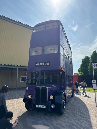 The image depicts the Knight Bus from the Harry Potter franchise on display in Nerima, Japan. The purple double-decker bus is parked on a brick-paved area. A red telephone box is behind the bus. Two people stand near the rear of the bus, one taking a photograph. Further back, two more individuals are walking away from the bus near a sign displaying the "KB" symbol and a London Underground sign. A low, corrugated-roofed building is visible on the left. The sky is light blue with some clouds.