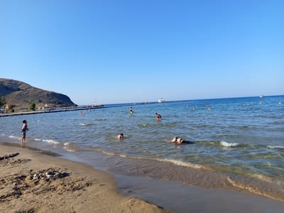 In Georgioupoli, Greece, a tranquil beach scene unfolds under a clear blue sky. The foreground is dominated by a sandy shoreline with a small arrangement of stones. Gentle waves lap at the wet sand, leading into the calm, shallow waters of the sea.
Several individuals are enjoying the water. Closer to the shore, a young person stands at the edge, looking out towards the sea. Further out, a person swims on their back, and another appears to be swimming or wading. In the mid-ground, more people are visible, some swimming, others standing in the water.
The seascape stretches to the horizon, where a long, dark pier extends into the sea. Along the pier, several figures can be seen, suggesting a public walkway or harbor access. A small white building, possibly a chapel, stands on the pier, marked by a Greek flag. To the left, a tree-covered hill rises from the coastline, forming a natural backdrop. A sailboat is visible in the distance, adding to the serene coastal atmosphere. The overall impression is one of a peaceful summer day at the beach.