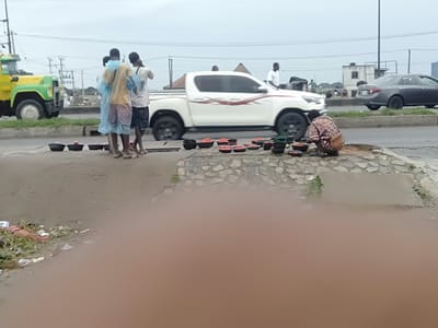 The image shows a roadside scene in Owode-Elede, Nigeria. A woman squats beside numerous bowls filled with a red produce, possibly tomatoes or peppers, laid out for sale on a concrete patch. Three men stand nearby, one wearing a blue plastic poncho. A white pickup truck passes on the road behind them, along with other vehicles. A green and yellow truck is parked farther away. Power lines stretch across the sky. Litter is scattered on the ground in the foreground. The sky is overcast.