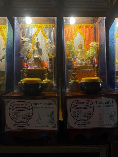The image displays two adjacent, illuminated devotional display cases, likely situated within a temple or shrine in Ban Hin Sam Kon, Thailand. Both cases are constructed with dark wooden frames and glass fronts, each containing a seated Buddha statue.
The left display case features a translucent or light-colored Buddha statue, centrally positioned on an ornate golden pedestal. The interior background consists of a shimmering silver fabric panel, topped with a golden-yellow fringed valance. Two bouquets of white, artificial flowers are placed symmetrically on either side of the statue. In front of the Buddha, two electric 'flame-effect' lamps, emitting an orange-red light, stand on golden bases. Below this case, on a dark wooden shelf, a black offering bowl with a yellow slotted lid is visible. A rectangular white sign with a red border is affixed to the shelf, bearing Thai script and the English text "NIGHT WEDNESDAY," along with graphic representations of currency and symbolic Buddhist imagery.
The right display case holds a golden Buddha statue, similarly seated on an elaborate golden pedestal. Its interior background is an orange-gold patterned fabric panel, also crowned with a golden-yellow fringed valance. Two bouquets of light green/yellow artificial flowers flank this statue. Identical electric 'flame-effect' lamps, glowing orange-red, are positioned in front. Below this case, a black offering bowl with a yellow slotted lid is present. An accompanying white sign with a red border displays Thai script and the English text "THURSDAY," alongside currency graphics and symbolic Buddhist figures, including a standing Buddha. The environment beyond the cases appears dimly lit.