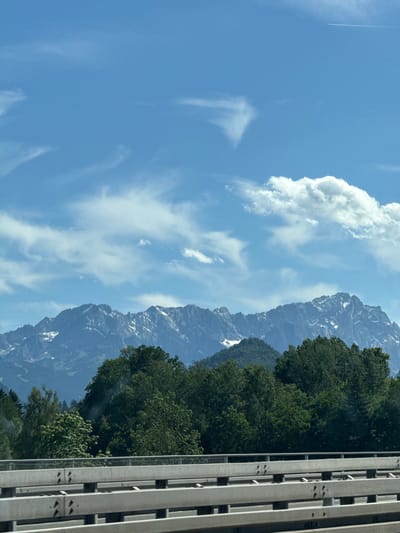 The image displays a scenic view of mountains and trees, with a clear blue sky dotted with white clouds. In the foreground, a concrete barrier with metal railings is visible, suggesting a road or bridge. The majestic, snow-capped mountains rise in the background, partially obscured by lush green trees. The overall impression is one of natural beauty and tranquility. The location of Ohlstadt, Germany is known for its picturesque landscapes, often featuring the Alps and rolling hills.