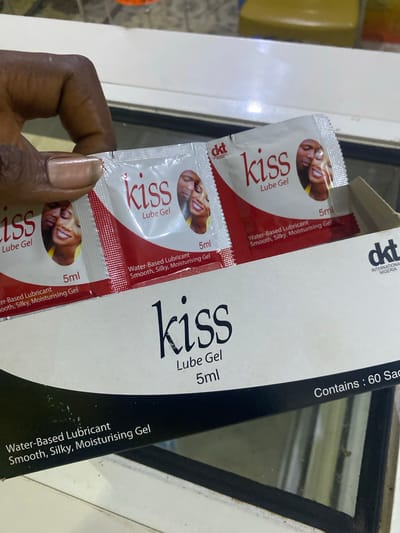 A hand with manicured nails holds three individual sachets of "Kiss Lube Gel" in Jere South, Nigeria. Each sachet is white with a red accent and features an image of a man and woman, the "Kiss" logo, and product details. The sachets are placed in a cardboard box, also labeled with "Kiss Lube Gel" and "DKT INTERNATIONAL NIGERIA." The background suggests an indoor setting, possibly a retail environment, with blurry yellow and white elements.