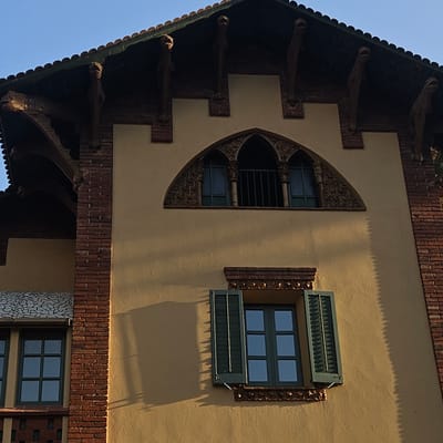 This is a close-up, low-angle shot of the upper facade of a building in Girona, Spain. The building exhibits distinctive architectural features with a mix of materials. The main wall is painted a warm, sandy yellow. Red brickwork is visible along the edges, forming pillars and decorative bands.
The roofline is steep and features dark tiles. Below the eaves, there are several ornate, carved wooden corbels that resemble stylized gargoyles or mythical creatures. These corbels protrude outwards, supporting the roof structure.
There are two windows visible in the frame. The upper window is a large, arched opening with intricate, dark, sculpted ornamentation around its perimeter and framing the glass panes. The window itself has multiple panes and is barred on the inside. The lower window is smaller and rectangular, with dark green shutters that are partially open, revealing a multi-paned window behind them. Decorative brickwork with a repeating pattern is present above and below this lower window.
The building's surface is uneven, with shadows cast by the architectural elements, creating a play of light and dark. The sky above is a clear, bright blue. The style of the architecture, particularly the sculpted corbels and the ornate window, suggests a modernist or Art Nouveau influence, common in cities like Girona that have a rich architectural history.