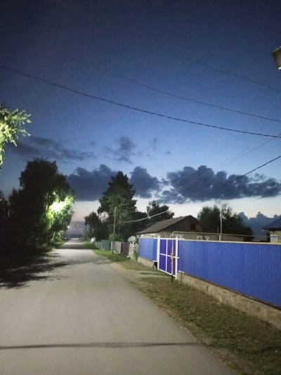 The image depicts a dimly lit rural road in the city of Voroshilovka, Kazakhstan, at twilight or night. The road is asphalt and appears to be empty of people or vehicles. On the right side of the road, a bright blue corrugated metal fence with white vertical supports runs along the property line of several houses. Behind the fence, parts of houses and trees are visible. The left side of the road is bordered by trees, with one tree illuminated by a bright light source, casting long shadows on the road. The sky above is a deep blue, with scattered dark clouds. Power lines are strung across the sky. A street light is visible on a pole in the upper right corner of the image. The overall atmosphere is quiet and still.