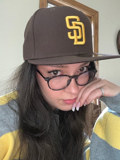 The image displays a person from the chest up, situated indoors, looking directly into the camera. The individual wears a dark brown baseball cap featuring a yellow "SD" logo on the front. Long, dark hair cascades over the shoulders. Eyeglasses with dark, possibly tortoiseshell-patterned frames are positioned on the face. The left hand, adorned with a silver-toned ring featuring multiple clear stones on the ring finger, rests with its long, square-tipped, French-manicured fingernails positioned near the chin and lower lip. The person is wearing a sweater characterized by horizontal stripes of light grey and yellow. The background consists of a plain, light-colored wall, with a partial view of a wooden door frame or window frame visible on the right side. The image contains no discernible features that would allow for a precise location identification within Glendale, United States.