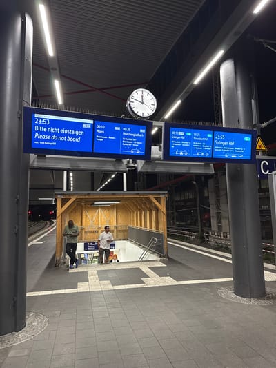 The image depicts a nighttime scene at a train station in Duisburg, Germany. The focal point is a large, illuminated digital display board showing train departure and arrival information. The board prominently features the text "Bitte nicht einsteigen / Please do not board" for a 23:53 departure from Bottrop Hbf. Other train information for various destinations and times is visible, including departures for Moers, Mönchengladbach, and Solingen Hbf.
In the foreground, two men stand on the platform. One man, wearing a green shirt and black pants, is looking at his phone. The other man, in a white t-shirt and shorts, is holding a beverage and also looking towards the display board. Behind them, a covered staircase leads down, with a sign indicating "1-4 und 8-" and "Ausgang City / Reisezentrum" (Exit City / Travel Center).
The platform itself is paved with dark tiles and marked with tactile paving for the visually impaired. Industrial elements like metal support columns, overhead lighting fixtures, and safety barriers are visible. The overall environment suggests a functional, modern train station operating at night.