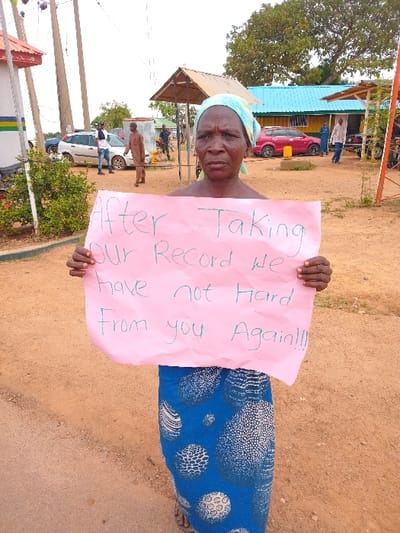 An older woman is standing in a dirt road and holding a pink sign in front of her. The sign reads "After Taking Our Record We Have Not Heard From You Again!!!" She is wearing a blue and white patterned skirt and a blue headscarf. There are two men and a car visible in the background. This scene likely takes place in Ungwan Tanko, Nigeria, which is located at the given coordinates.