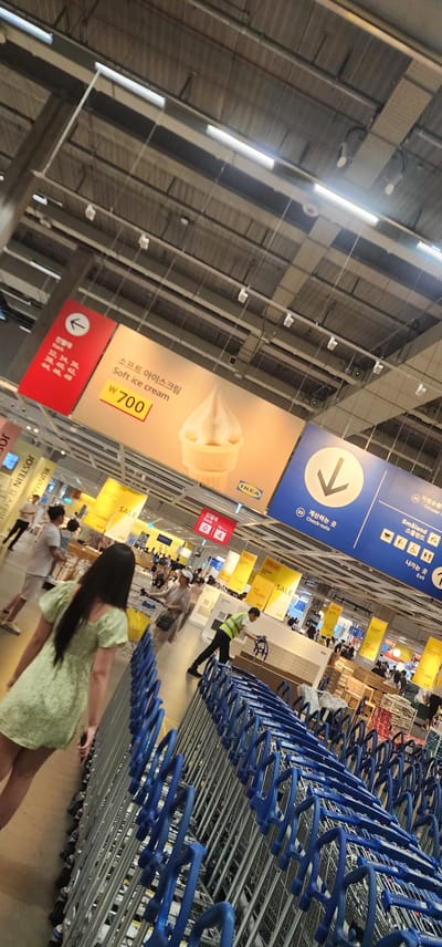 The image shows the interior of a large retail store, likely an IKEA, in Gwangmyeong, South Korea. The scene is dominated by rows of blue and silver shopping carts in the foreground, with a woman in a light green dress walking away from the camera. In the background, there are various signs and displays. A prominent sign above features an image of soft-serve ice cream with text in Korean and English, indicating "Soft ice cream" and a price of "₩ 700". Another sign with a downward-pointing arrow and the text "Check-outs" in English and Korean is visible. Other signs indicate "SALE" in yellow. Several people are browsing and shopping in the background, and overhead lighting and exposed ceiling structures are visible. The overall impression is a busy retail environment.