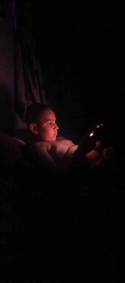 The image depicts a young person, likely a male, in a dark room, illuminated by a reddish glow from an unseen source. The person is shown in profile, with their head tilted downwards, and their gaze directed towards something held in their hands. Their face is partially illuminated, highlighting their features with a warm light. The surroundings are obscured by darkness, with only a hint of fabric or drapery visible in the background to the left. The overall atmosphere is intimate and focused, suggesting a private moment of engagement with the light source. There is no discernible urban environment or specific location in Khartsyzk, Ukraine identifiable within the frame.