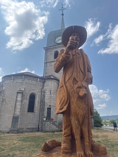 An outdoor, eye-level, full-shot photograph features a large, intricately carved wooden sculpture of a man wearing a wide-brimmed hat and a long coat, holding a glass in his right hand. A wolf is carved and integrated into the lower portion of the sculpture, appearing to be part of the man's cloak. The sculpture stands in a grassy area in front of a stone church with a steeple and a clock face. The sky above is blue with scattered white clouds. In the background, a paved path leads away from the church, with a person walking in the distance. The location is identified as Grande-Rivière Château, France.